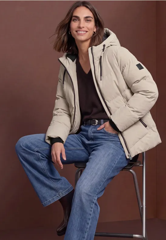 Frau trägt beige Steppjacke, schwarzen Pullover und Jeans, sitzt auf Hocker.