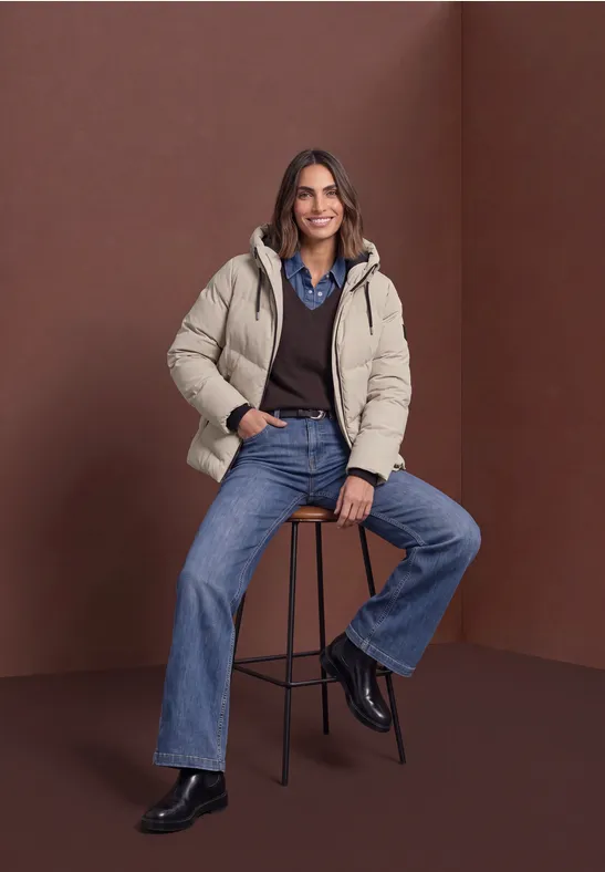 Frau in beiger Steppjacke, Hemd und Jeans sitzt lässig auf einem Hocker vor braunem Hintergrund.
