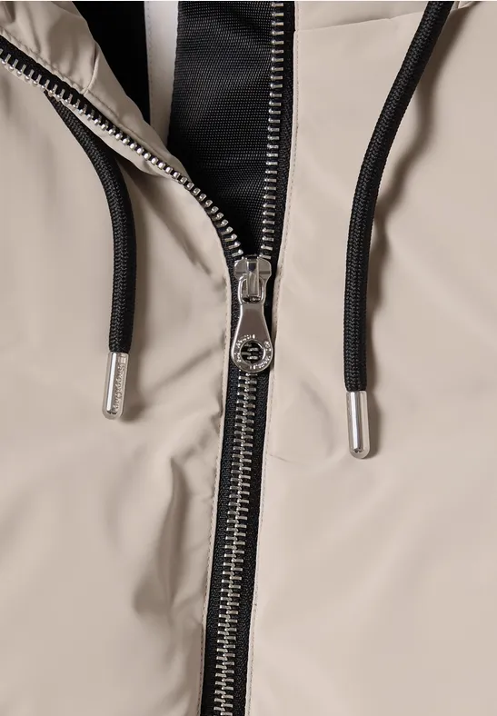Nahaufnahme eines Reißverschlusses an einer beigen Jacke mit schwarzem Kapuzenfutter und Kordeln.
