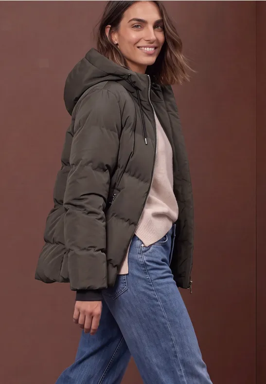 Lächelnde Frau in gesteppter Kapuzenjacke, hellem Pullover und Jeans.