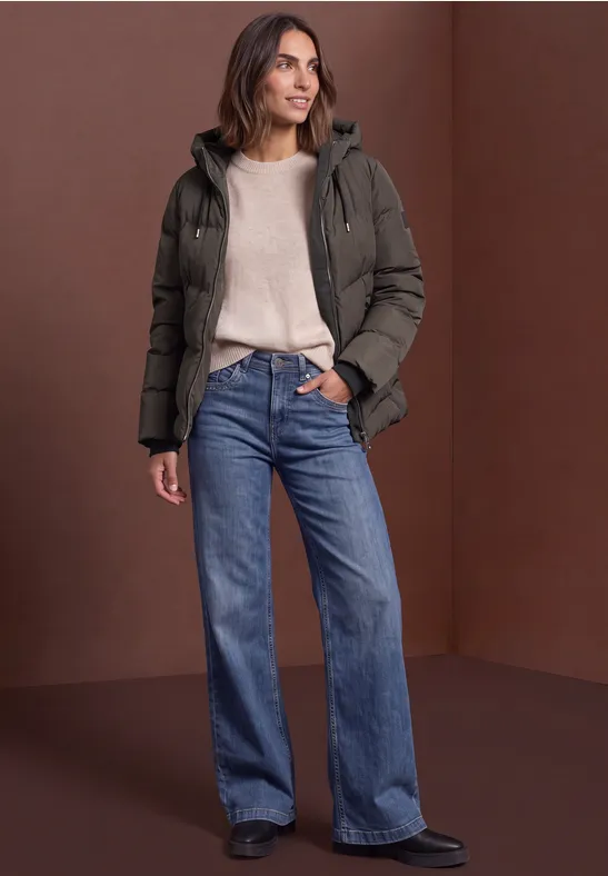 Frau trägt grüne Steppjacke, beigen Pullover und weite blaue Jeans vor braunem Hintergrund.