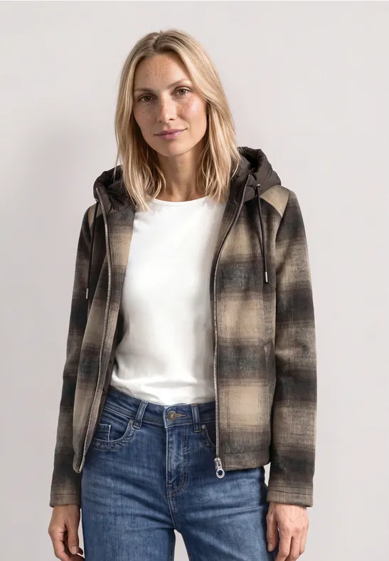 Frau mit blonder Mähne trägt eine karierte Jacke mit Kapuze, weißes Shirt und blaue Jeans.