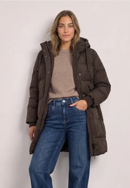 Frau in langer dunkelbrauner Steppjacke mit Jeans und Pullover, entspannt lächelnd