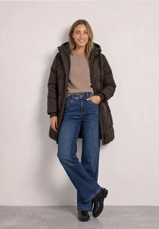 Junge Frau in braunem Steppmantel, beigem Pullover und Jeans vor neutralem Hintergrund, lächelnd.