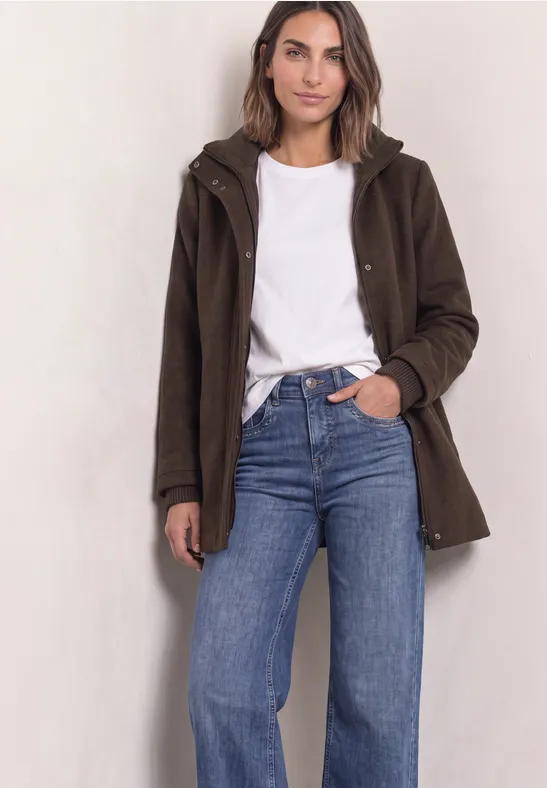 Frau in weißem Shirt, blauer Jeans und braunem Mantel vor hellem Hintergrund