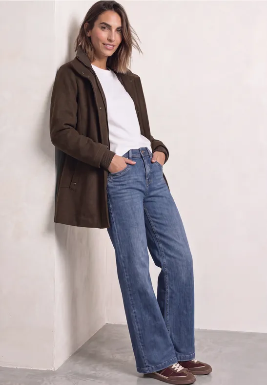 Eine Frau lehnt an einer Wand und trägt einen braunen Mantel, weißes Shirt, blaue Jeans und braune Schuhe.