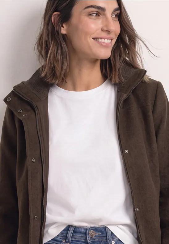 Frau mit schulterlangem braunem Haar, lächelnd, trägt weißes Shirt und olivgrüne Jacke.