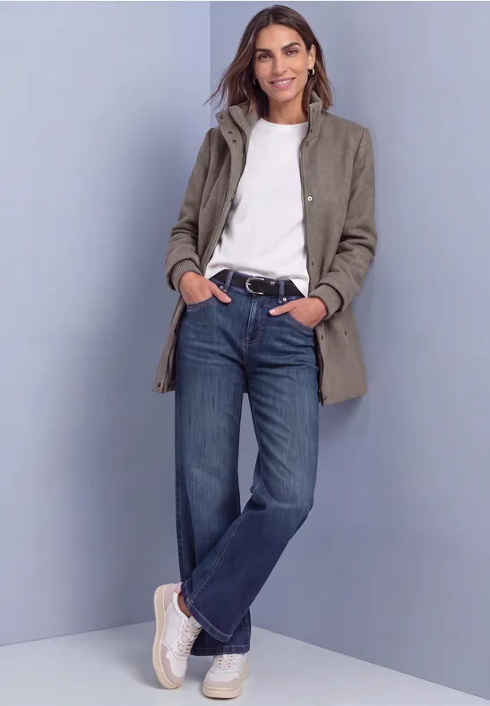 Frau in grauem Mantel, weißem Shirt, blauen Jeans und weißen Sneakern steht entspannt, lehnt an Wand.