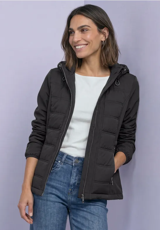 Frau in schwarzer Steppjacke mit Kapuze, weißem Shirt und blauen Jeans, lächelnd.