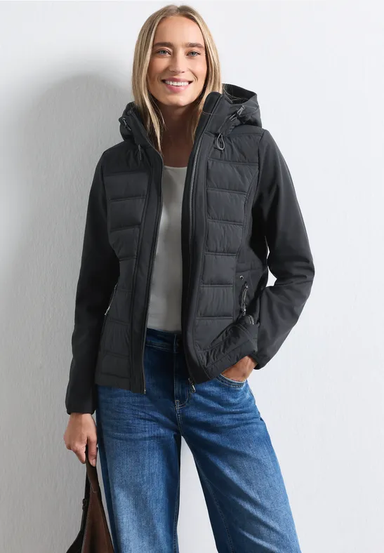 Lächelnde Frau in schwarzer Kapuzen-Steppjacke, blauer Jeans und Tasche vor weißem Hintergrund.