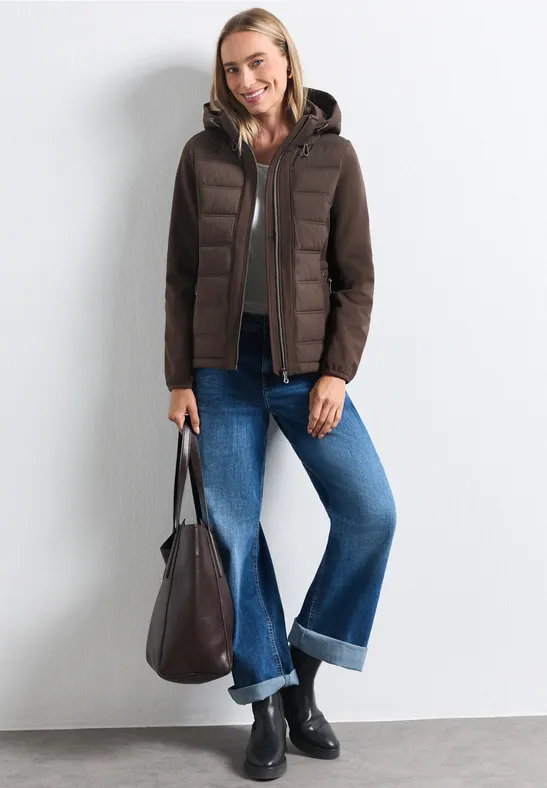 Frau in brauner Steppjacke, weitem Jeans, schwarzen Stiefeletten und Tasche vor weißer Wand.