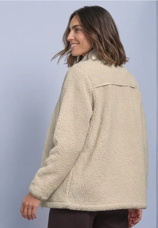 Frau in beige flauschiger Jacke vor neutralem Hintergrund, halb gewandt, lächelt.