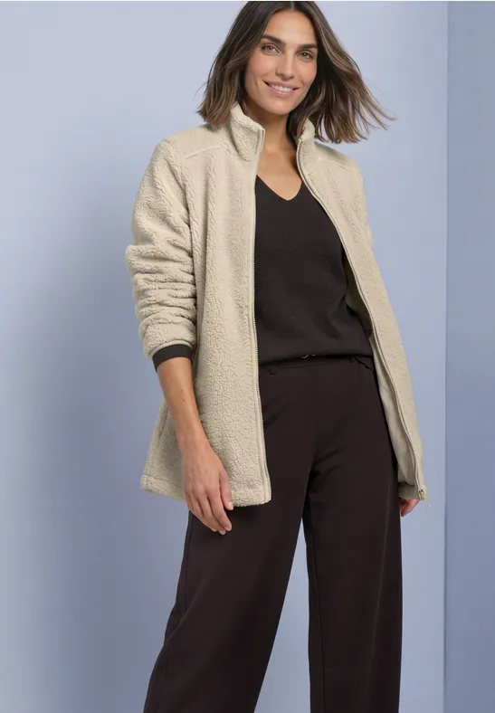 Eine Frau trägt eine beige Teddyjacke, schwarzes Top und Hose, sie lächelt vor hellem Hintergrund.
