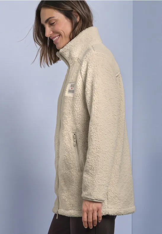 Eine Frau trägt lächelnd eine hellbeige Teddy-Fleecejacke mit Stehkragen vor hellem Hintergrund.