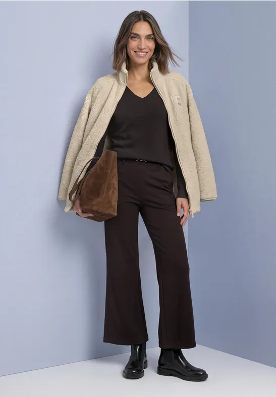 Frau vor blauem Hintergrund trägt beige Jacke, schwarzes Top, braune Hose, schwarze Stiefel und Handtasche.