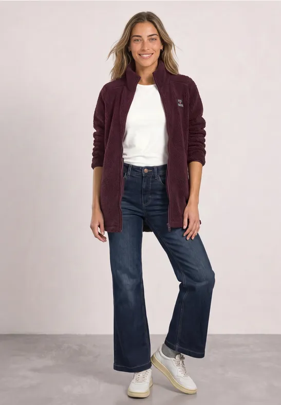 Une femme souriante porte une veste polaire bordeaux, un t-shirt blanc, un jean large et des baskets blanches.