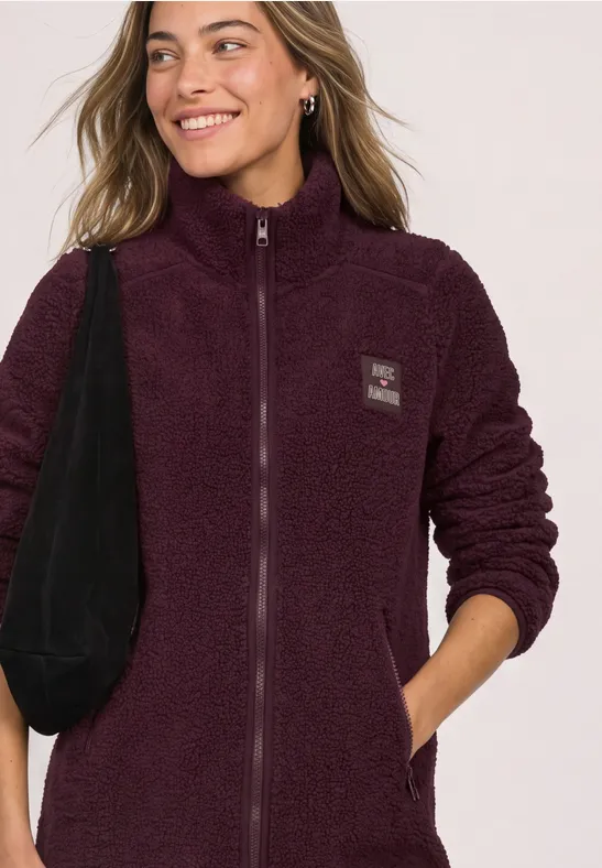 Une femme sourit détendue, portant une veste polaire prune zippée et un sac noir en bandoulière.