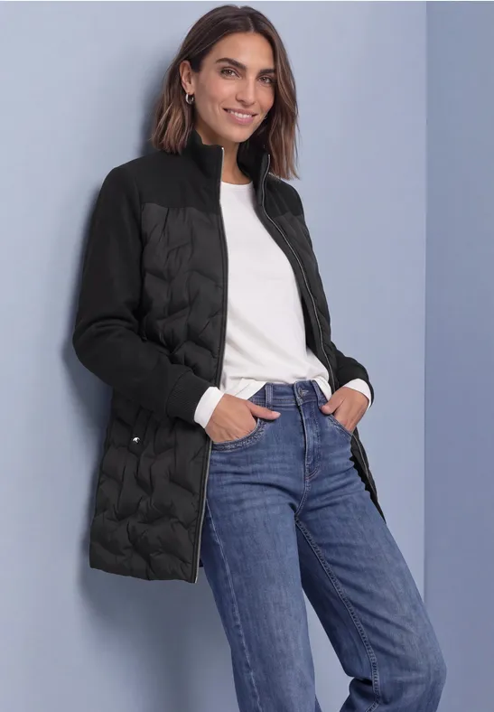 Frau in schwarzer Steppjacke, weißem Oberteil und blauen Jeans, lächelnd mit Händen in den Taschen vor grauem Hintergrund.