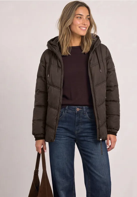 Frau trägt schwarze Daunenjacke, schwarzen Pullover, blaue Jeans und hält braune Tasche.