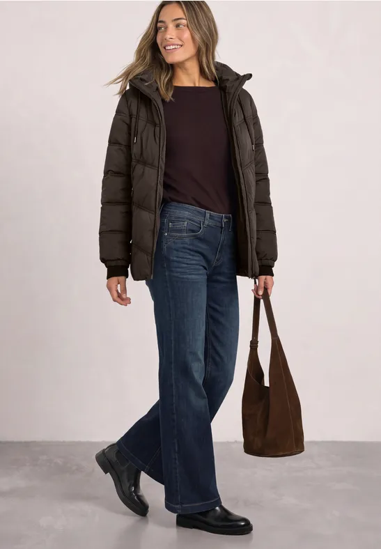 Frau in brauner Daunenjacke, dunklem Shirt, weiter Jeans und schwarzen Schuhen mit brauner Tasche.