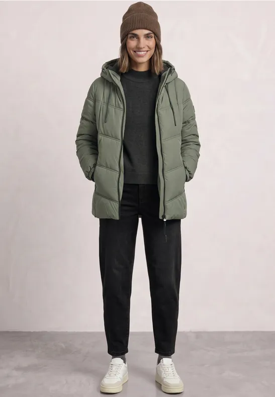 Eine Frau in grüner Steppjacke, schwarzer Hose, Mütze und weißen Sneakers steht vor hellem Hintergrund.