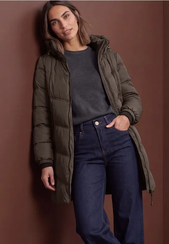 Frau in olivgrünem Daunenmantel, dunklem Pullover und Jeans vor braunem Hintergrund.