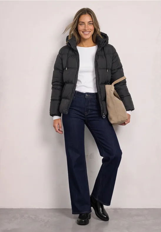 Eine lächelnde Frau trägt eine schwarze Steppjacke, ein weißes Shirt, weite Jeans und eine Tasche.