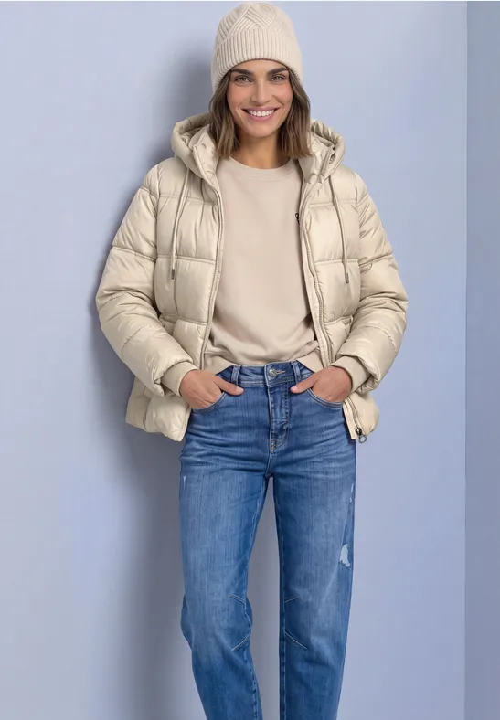 Eine Frau lächelt vor hellem Hintergrund, trägt eine beige Daunenjacke, Strickmütze, Pullover und blaue Jeans.