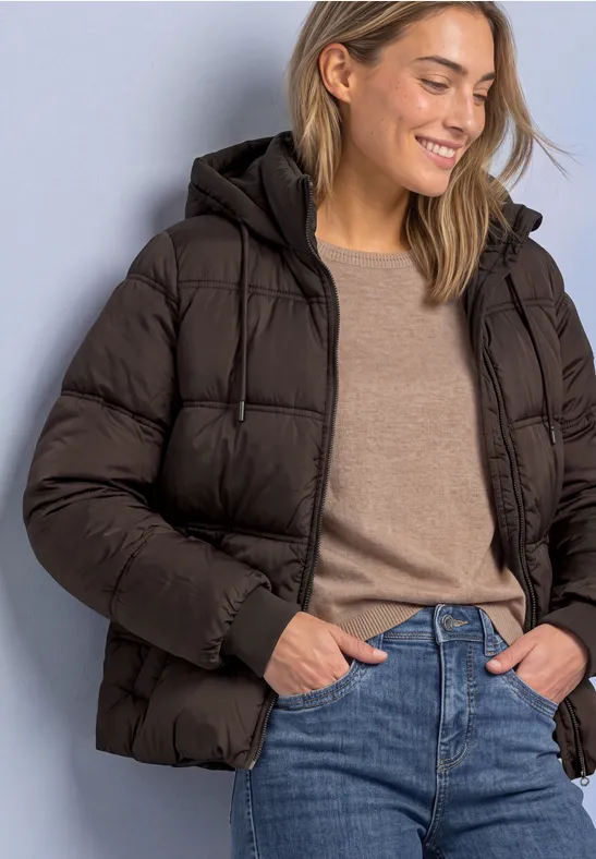 Junge Frau in dunkelbrauner Steppjacke mit Kapuze, Pullover und Jeans, lächelnd mit Händen in den Taschen.