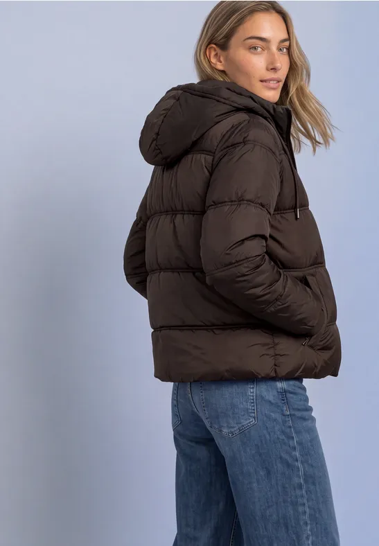 Eine Frau in brauner Steppjacke mit Kapuze und blauen Jeans vor hellem Hintergrund.