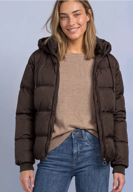 Eine lächelnde Frau in brauner Steppjacke, beigem Pullover und Jeans vor neutralem Hintergrund.
