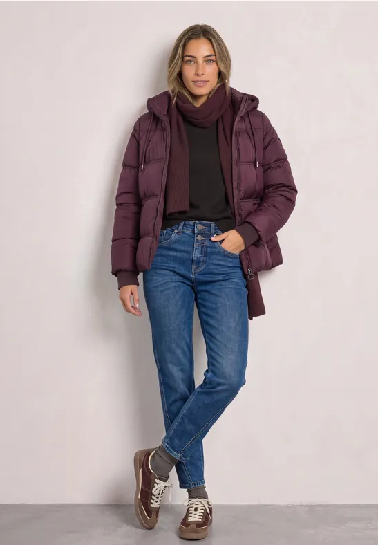 Frau in violetter Steppjacke, dunklem Pullover, blauen Jeans und Sneakern steht lässig vor einer Wand.
