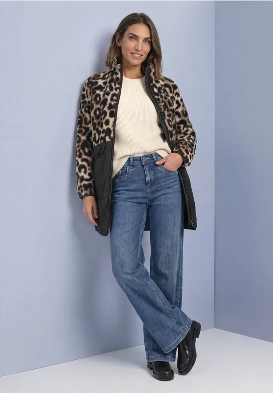 Frau in Leopardenmuster-Mantel, hellem Pullover und Jeans vor blauem Hintergrund.