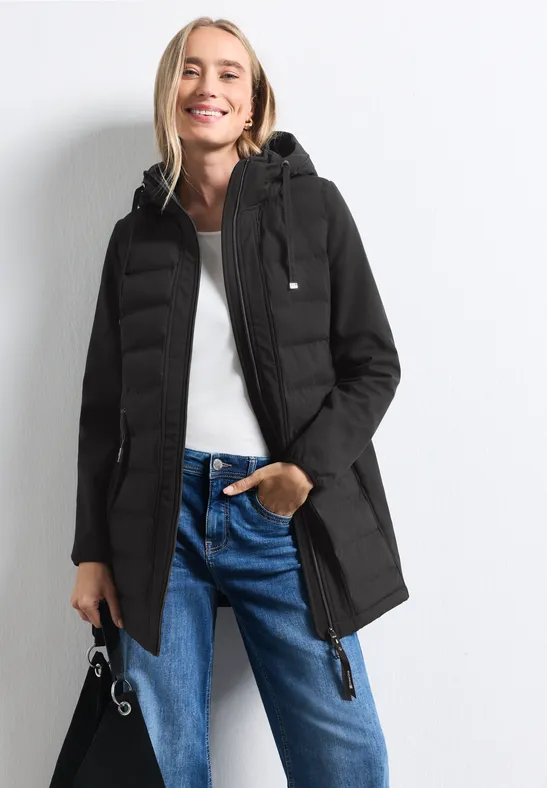 Frau trägt schwarzen Parka mit Kapuze, weiße Bluse und blaue Jeans, schaut lächelnd in die Kamera.