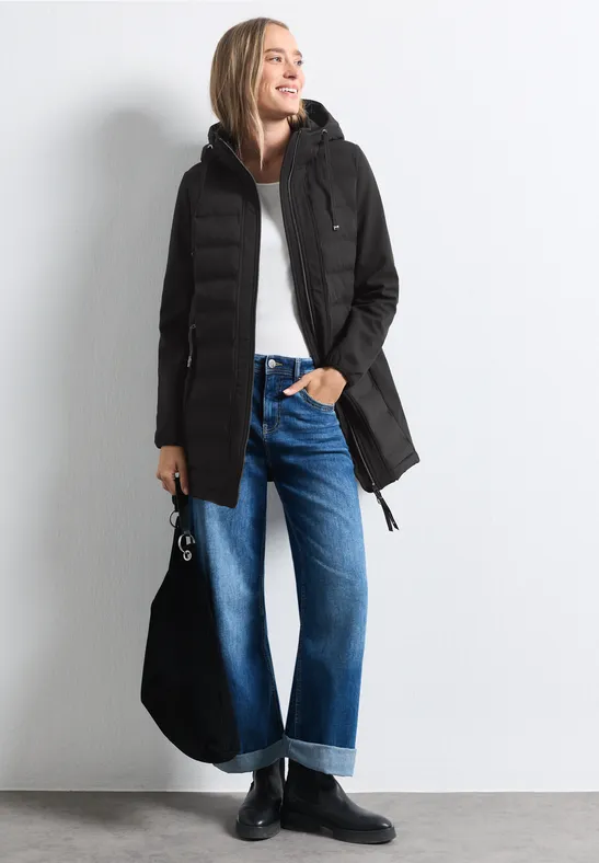Eine Frau in schwarzer Steppjacke, weißem Oberteil, weitem Jeans und schwarzen Stiefeln, mit Tasche an der Seite.