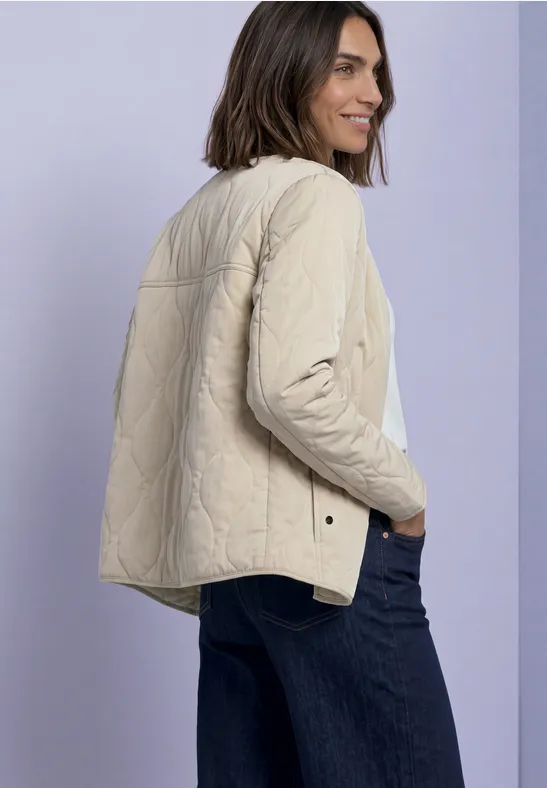Vrouw met halflang haar draagt een beige doorgestikte jas en blauwe jeans, staand voor een lichtgekleurde achtergrond.