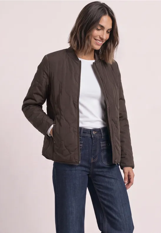 Frau trägt eine Steppjacke in Dunkelbraun, dazu weißes Shirt und Jeans, sie lächelt.