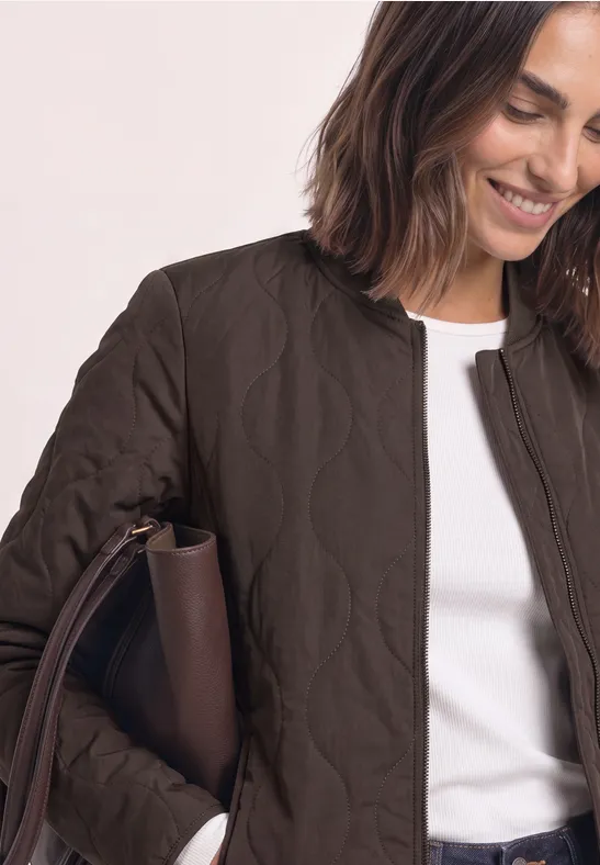 Frau mit brauner gesteppter Jacke und weißem Shirt, lächelnd und hält eine braune Handtasche.