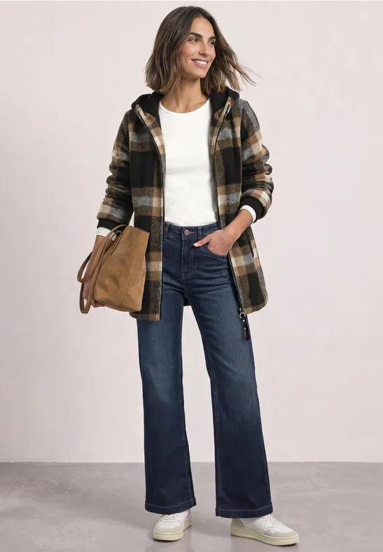 Frau in Jeans, weißem T-Shirt, kariertem Mantel und weißen Sneakern, lächelnd und mit Tasche.