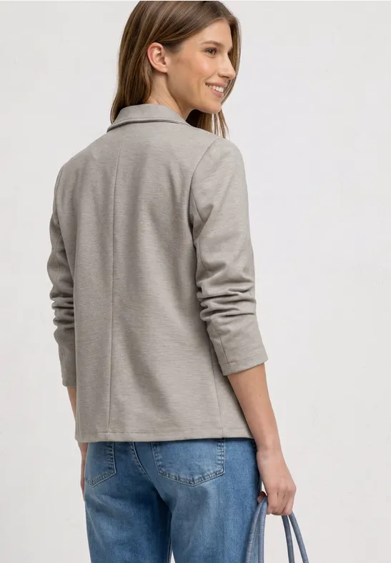 Een persoon gezien van achteren draagt een lichtgrijs jasje, blauwe jeans en houdt een tas vast.
