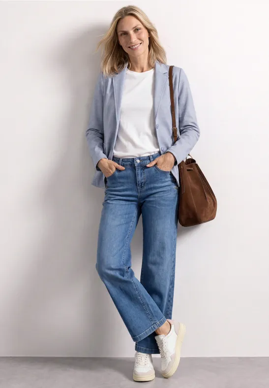 Leicht verschwommene Frau an der Wand, hellblauer Blazer, weißes Shirt, gerade Jeans, weiße Sneaker, braune Tasche.