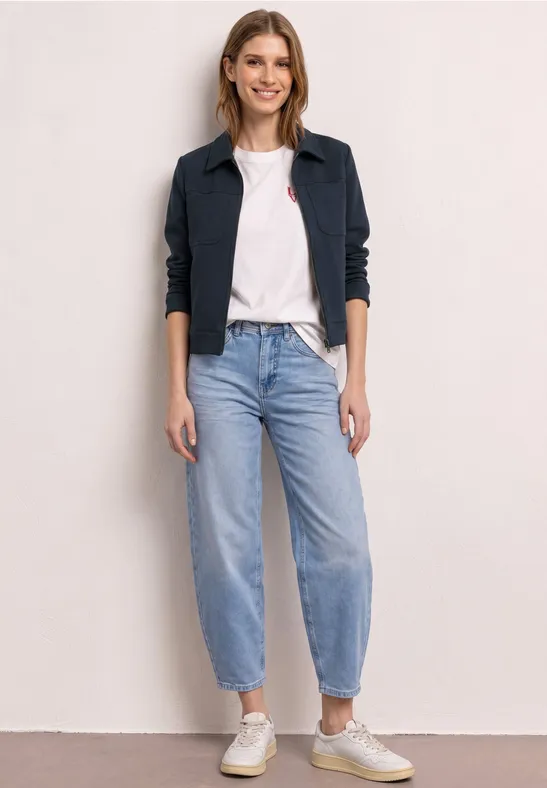 Verschleierte Person in weißem T-Shirt, dunkelblauer Jacke, hellblauen Jeans und beigen Sneakern vor heller Wand.