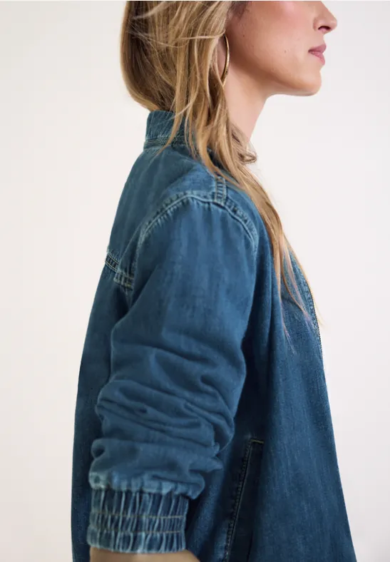 Zijaanzicht van een vrouw met lang blond haar en een spijkerjas tegen een lichte achtergrond.