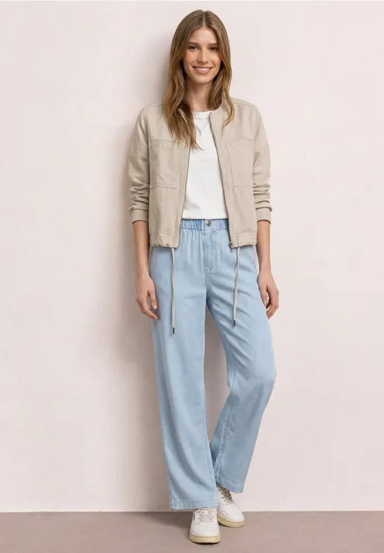 Vrouw met een beige leren jasje, wit shirt, ruime blauwe jeans en witte sneakers voor een lichte muur.