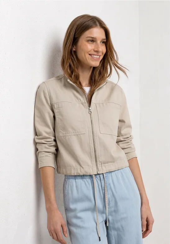 Een vrouw voor een witte achtergrond draagt een zandkleurig jasje met rits en een losse lichtblauwe broek.
