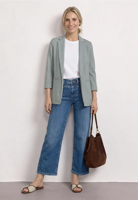 Frau in weißem T-Shirt, grauem Blazer, weiten Jeans, Sandalen und brauner Tasche.