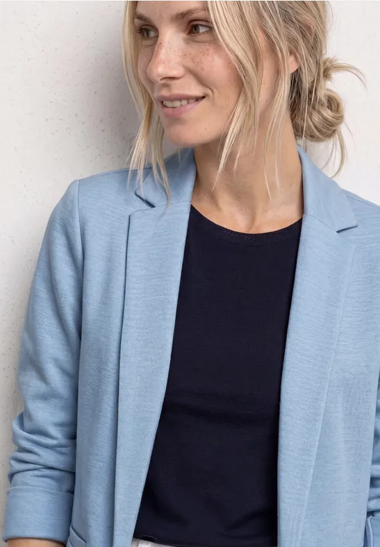 Femme au chignon blond porte un blazer bleu clair et un top noir sur fond clair.
