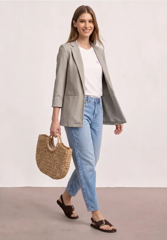 Femme portant un blazer gris, un t-shirt blanc, un jean bleu, sandales noires et tenant un sac en paille.