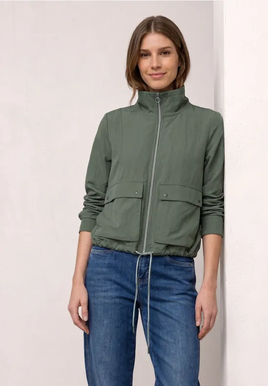 Frau in grüner Steppjacke und blauen Jeans vor hellem Hintergrund.