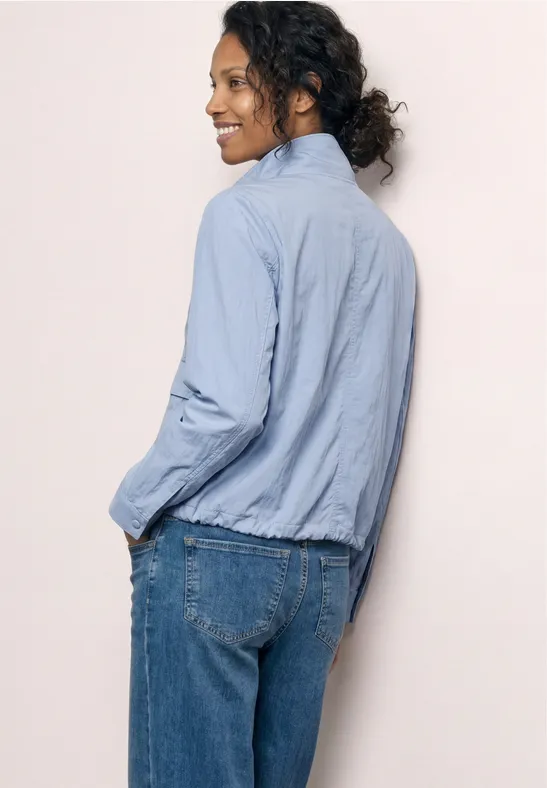 Personne de dos portant un chemisier bleu clair et un jean, cheveux bruns bouclés attachés, fond clair.